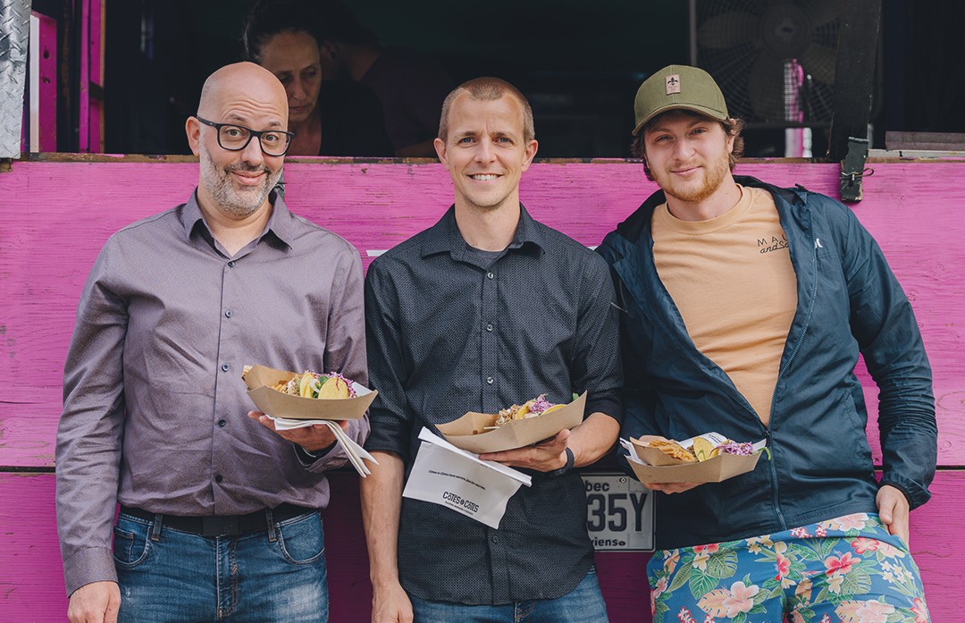 Trois hommes sourient devant un camion-restaurant rose, chacun tenant une assiette en carton de nourriture