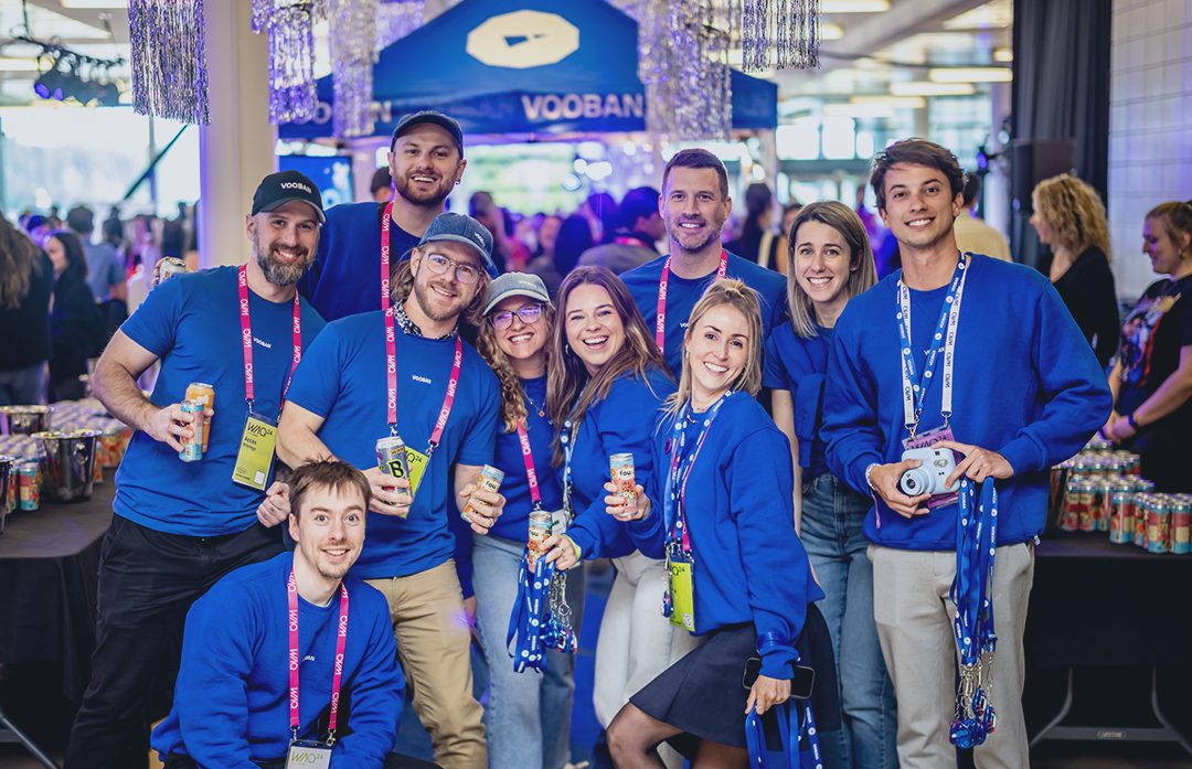 Des membres de l’équipe en chandail bleu sourient, canette en main, devant un kiosque Vooban lors d’un événement