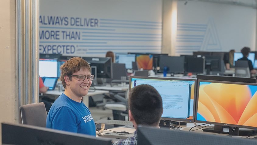 Un employé en t-shirt Vooban sourit dans un bureau ouvert, devant un mur « Always deliver more than expected »