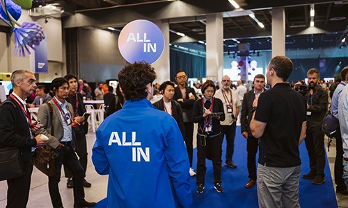 Visitors standing in an exhibition hall, one seen from behind in a blue jacket reading “ALL IN”