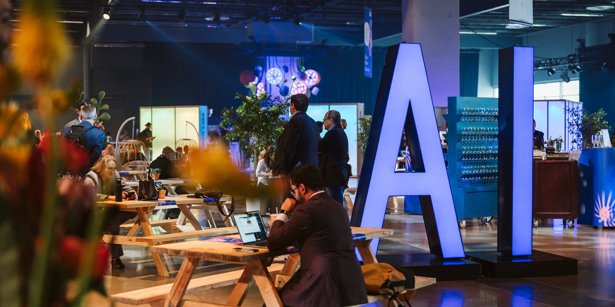 Grandes lettres lumineuses « AI » dans un salon, avec des personnes assises à des tables de pique-nique
