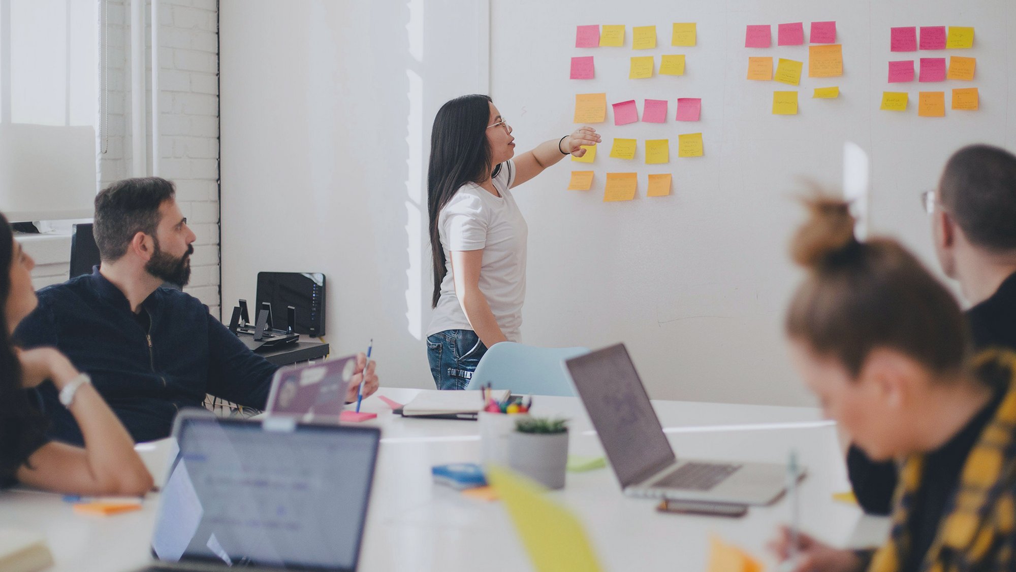 A woman points at sticky notes on a wall in front of colleagues seated with laptops