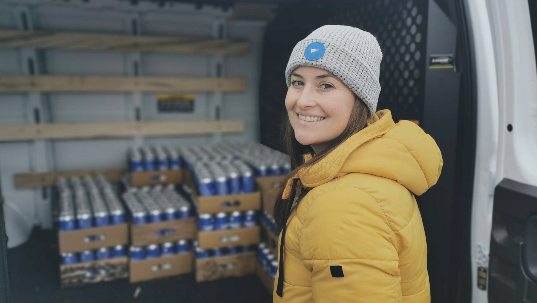 A smiling employee in a beanie and yellow coat in front of a van loaded with crates of cans