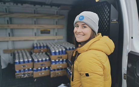 A smiling employee in a beanie and yellow coat in front of a van loaded with crates of cans