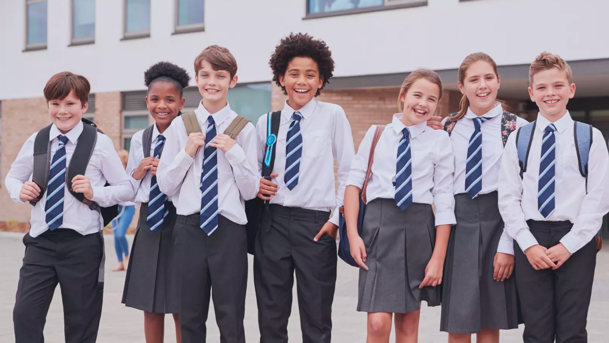 Seven students in school uniforms with backpacks pose in front of a building