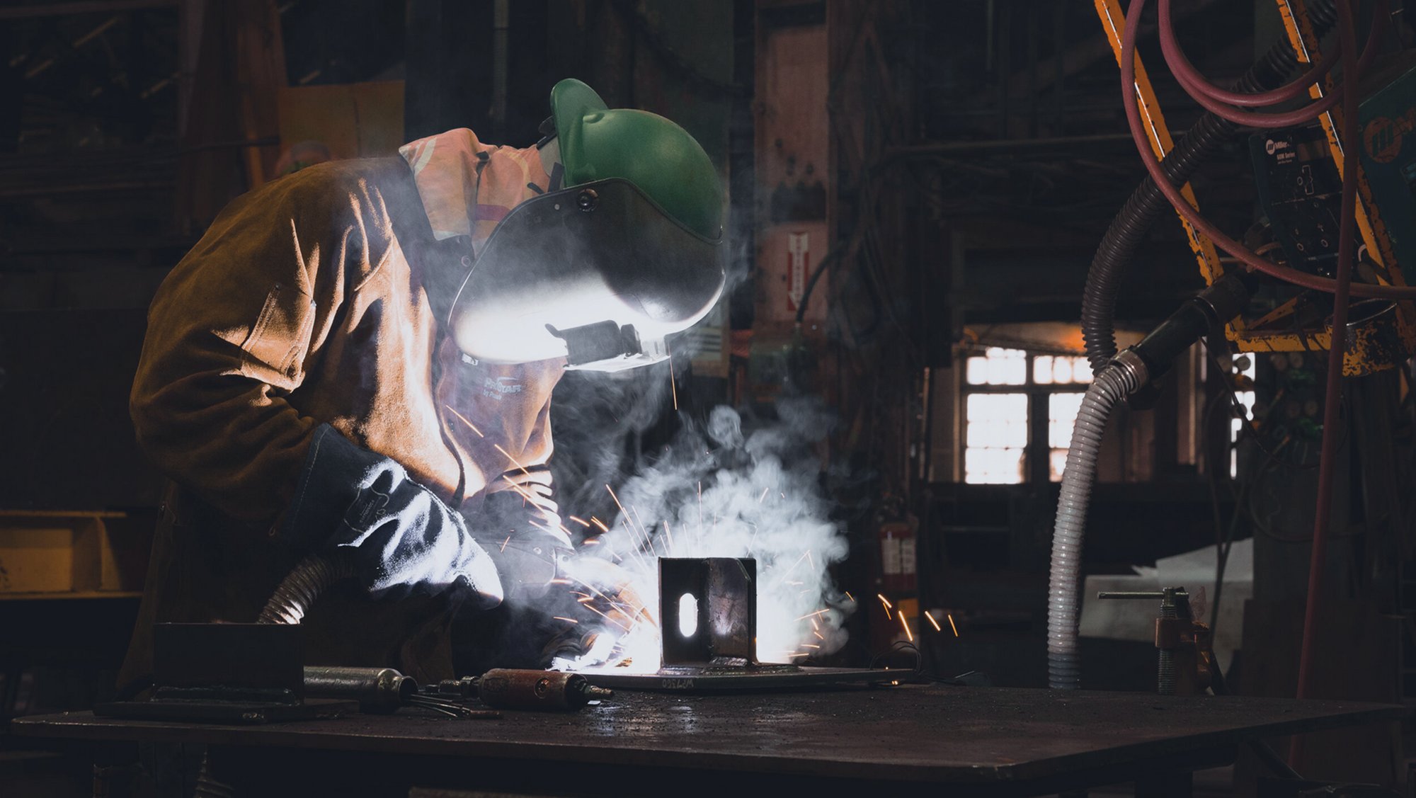 Welder in a helmet working on a steel part in a workshop, with sparks and smoke